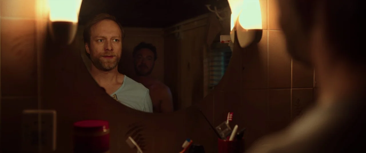 Speak No Evil (2022) movie still: medium — A dimly lit bathroom features brown tiled walls and two wall-mounted light fixtures. A man in a blue…