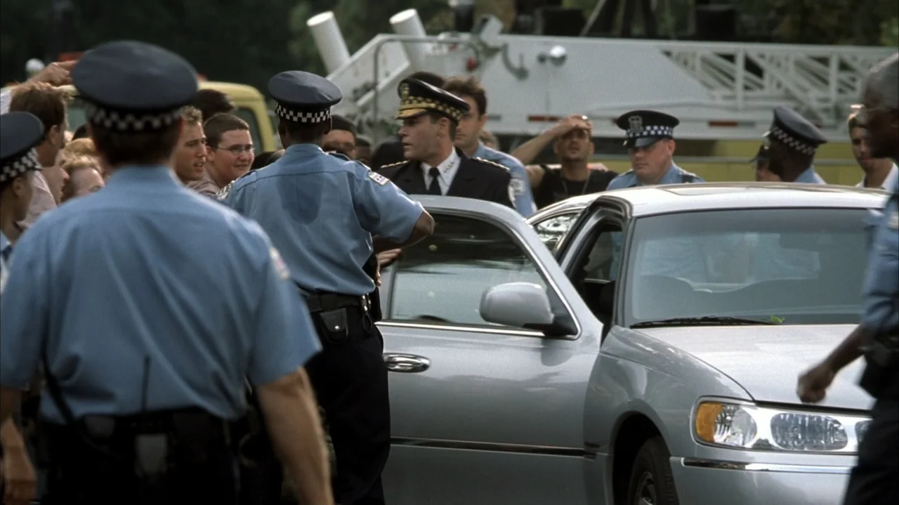 John Q (2002) movie still: medium — Multiple uniformed police officers gather around a silver sedan on a city road. The high-ranking officer in a…