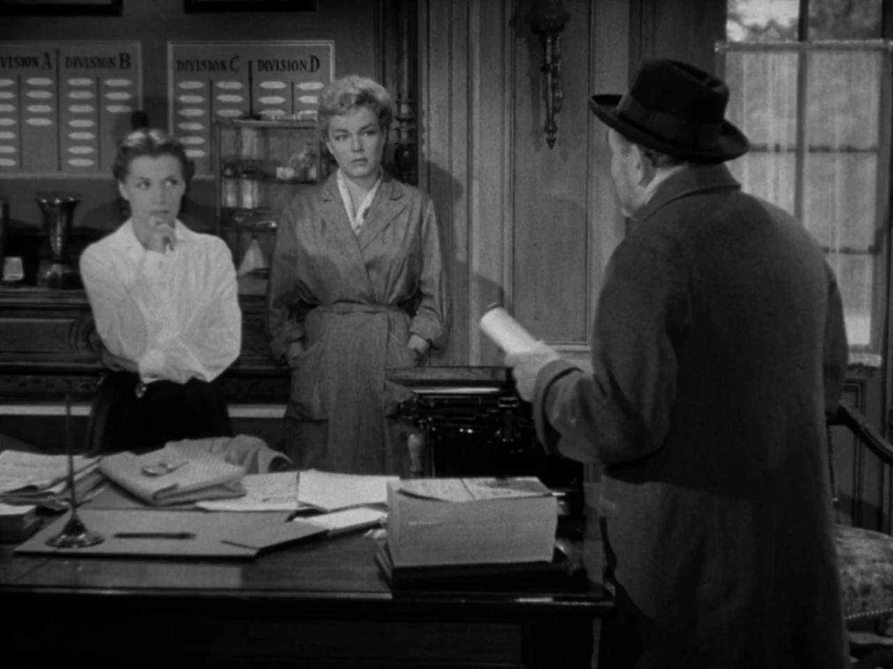 Diabolique (1955) movie still: medium — A wood-paneled office contains a desk with papers, a typewriter, and organization boards. Two women watch a…
