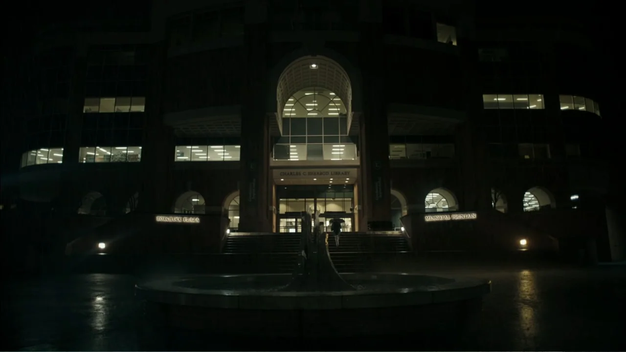 Alabama Snake (2020) movie still: wide — A modern brick university library building with a central fountain in the foreground. A single figure walks…