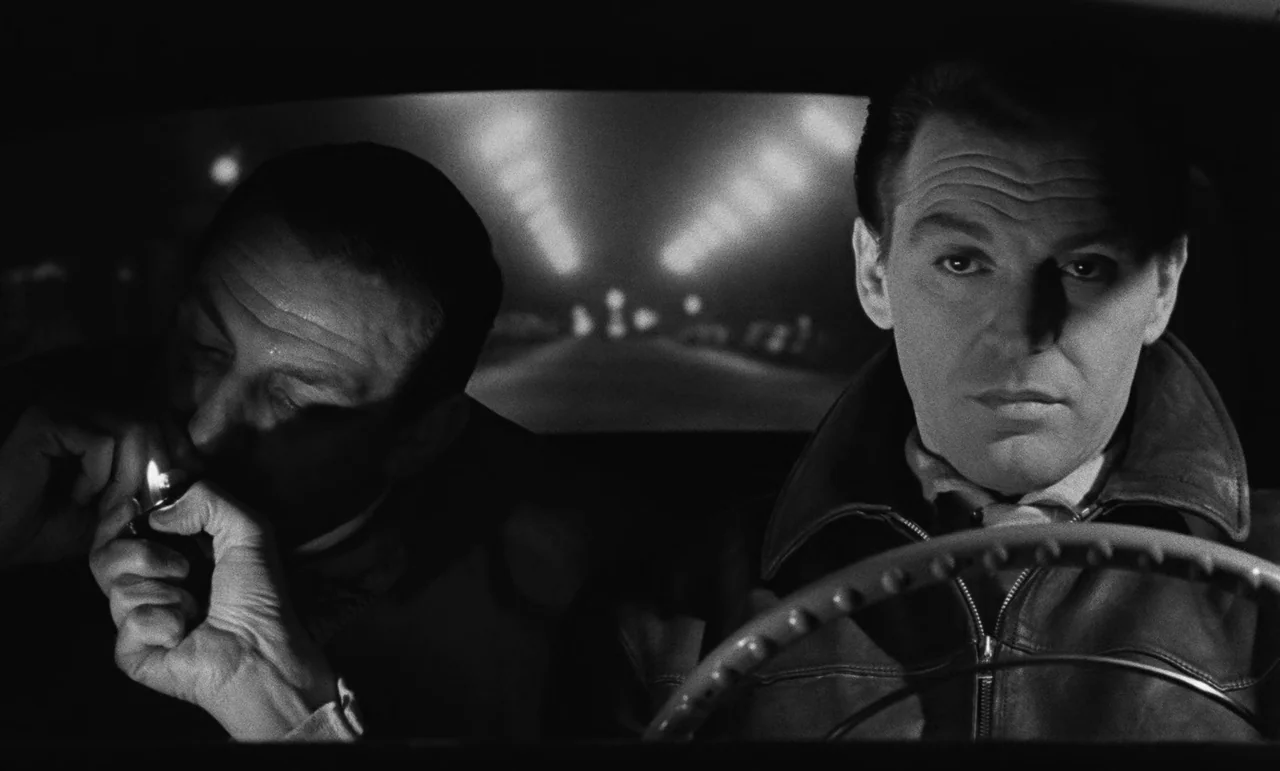 Witness In The City (1959) movie still: medium — Two men sit inside the front cabin of a vehicle at night. A long road with blurred streetlights is…