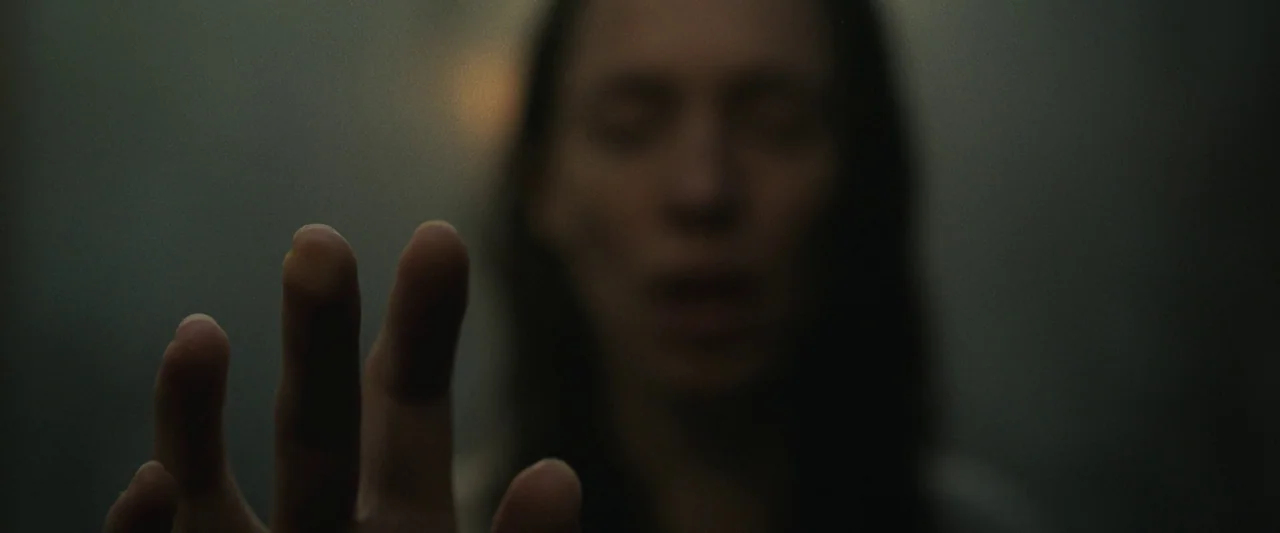 The Night House (2020) movie still: closeup — A dimly lit interior space features a transparent barrier between the viewer and subject. A woman…