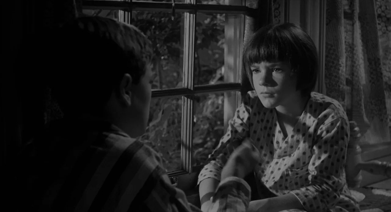 To Kill A Mockingbird (1962) movie still: medium — Two children sit near a wooden window frame in a dimly lit space. They face each other while…
