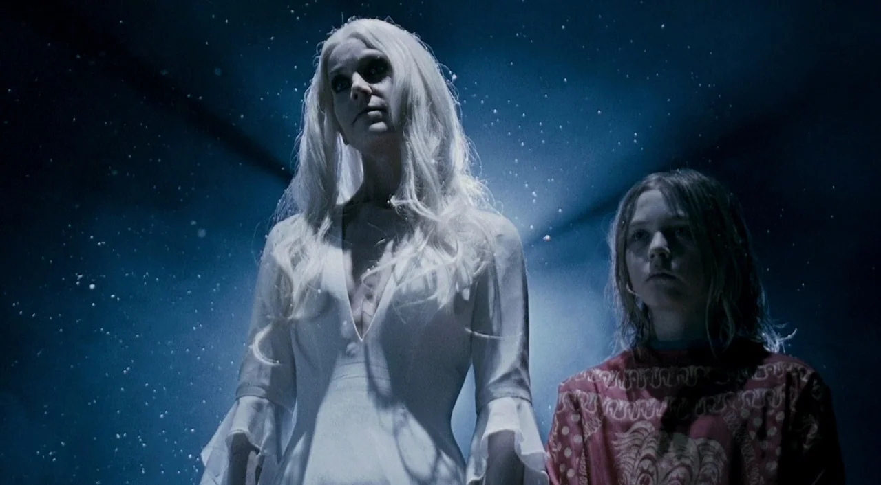 Halloween 2 (2009) movie still: medium — Two individuals stand against a dark background illuminated by floating white particles. A pale woman in a…