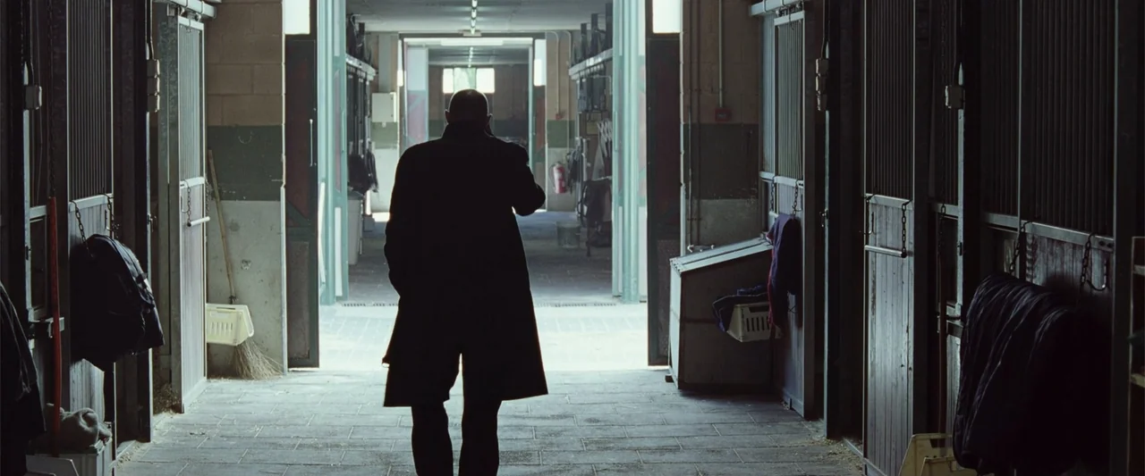 Anna (2013) movie still: fullbody — A long stable aisle features rows of wooden stalls with metal barred gates. A bald man wearing a long coat walks…