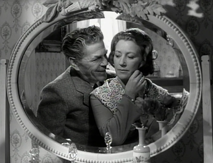 The Devil’s Eye (1960) movie still: medium — A domestic space features an oval vanity mirror reflecting an older couple. The man leans over the…