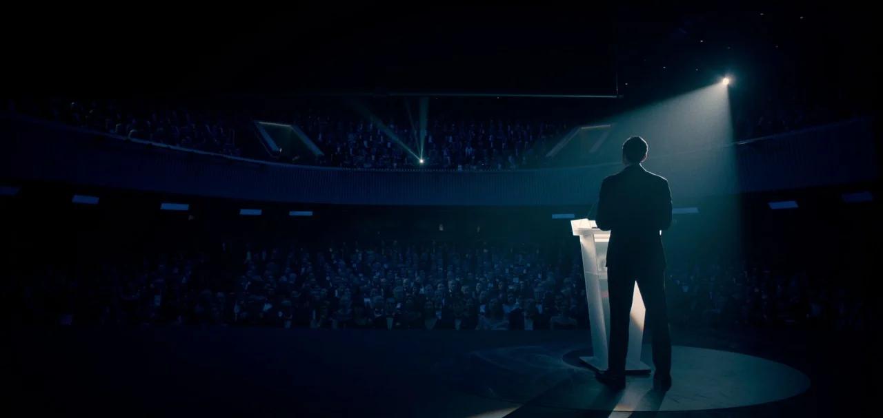 The Midnight Sky (2020) movie still: wide — A dark auditorium holds a large seated audience watching a solitary speaker. A man in a suit stands…