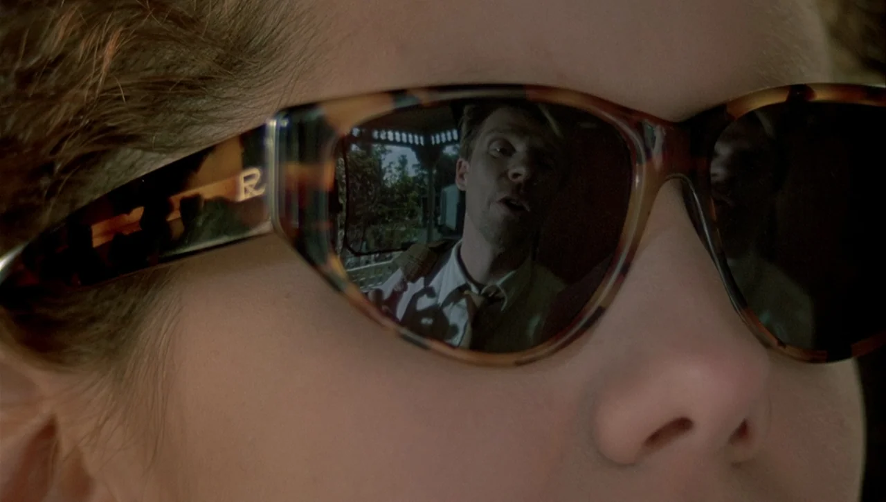 Necronomicon: Book of Dead (1993) movie still: closeup — A man in a white shirt and tie is seen reflected in a pair of glasses. The background shows…