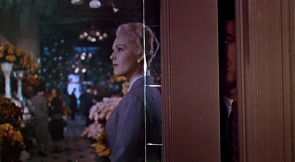 Vertigo (1958) movie still: medium — A flower shop interior with shelves of chrysanthemums and people in the background. A woman in a suit stands in…