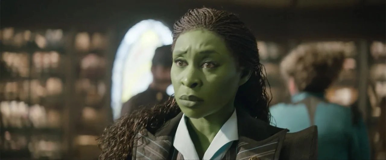 Wicked (2024) movie still: closeup — A dimly lit interior passenger cabin features a blurred arched window in the background. A woman with green…