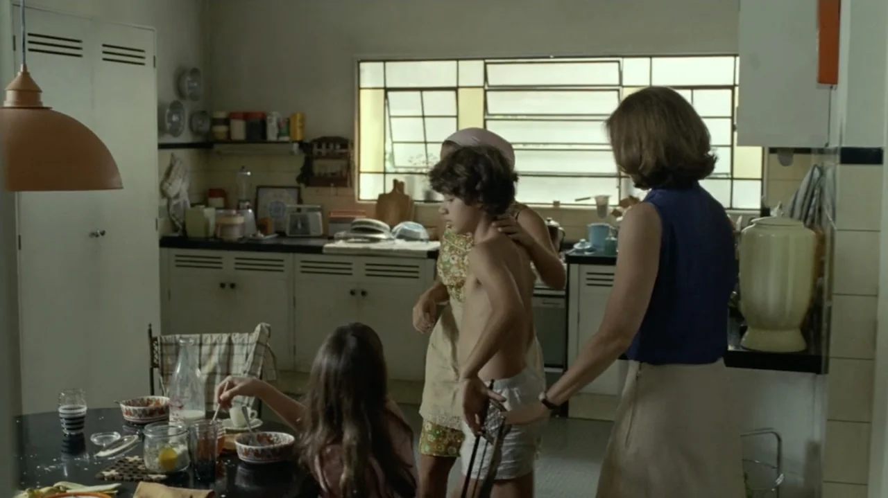 I'm Still Here (2024) movie still: medium — A 1970s interior with retro cabinetry and a large window above the counter. Three people interact in the…