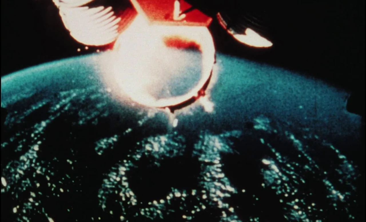 Sans Soleil (1983) movie still: establishing — A dark, grain-textured view of a planet from high orbit in space. A bright white thruster plume glows…