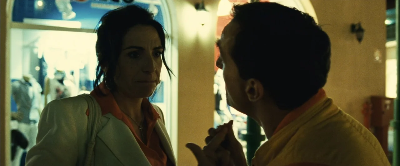 Reality (2023) movie still: medium — A commercial storefront exterior with large glass windows at night. A man gestures while talking to a woman…