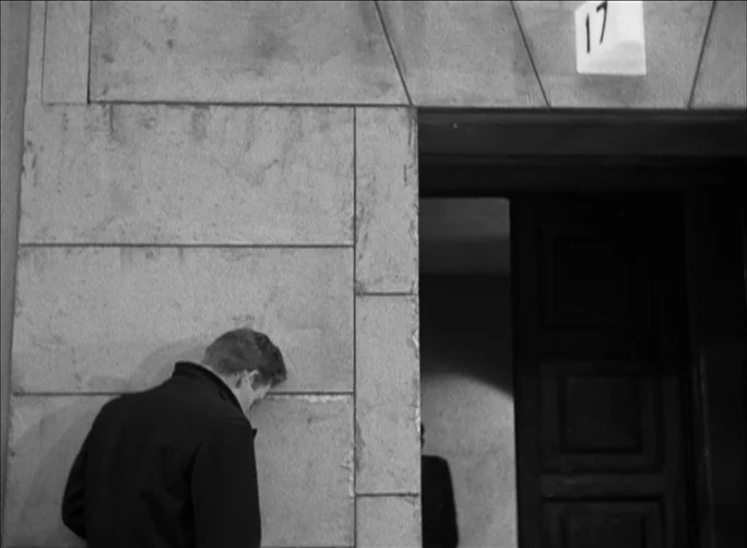 Summer with Monika (1953) movie still: medium — A stone building wall with a doorway marked with the number seventeen. A man in a dark coat stands…
