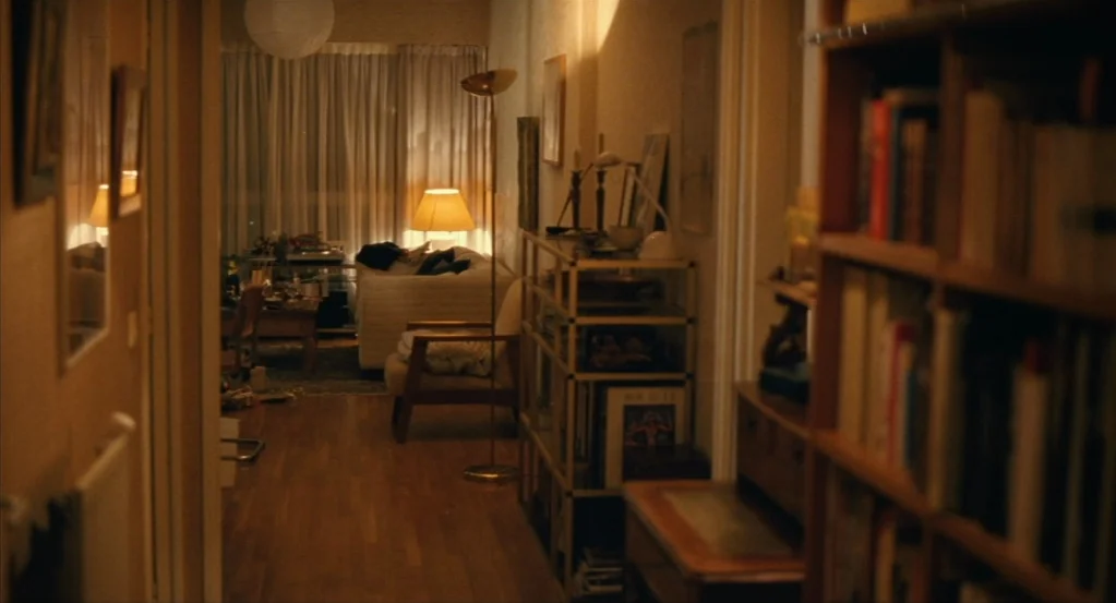 Things to Come (2016) movie still: wide — A dimly lit apartment interior features a hallway opening into a cozy lounge area. A tall floor lamp…
