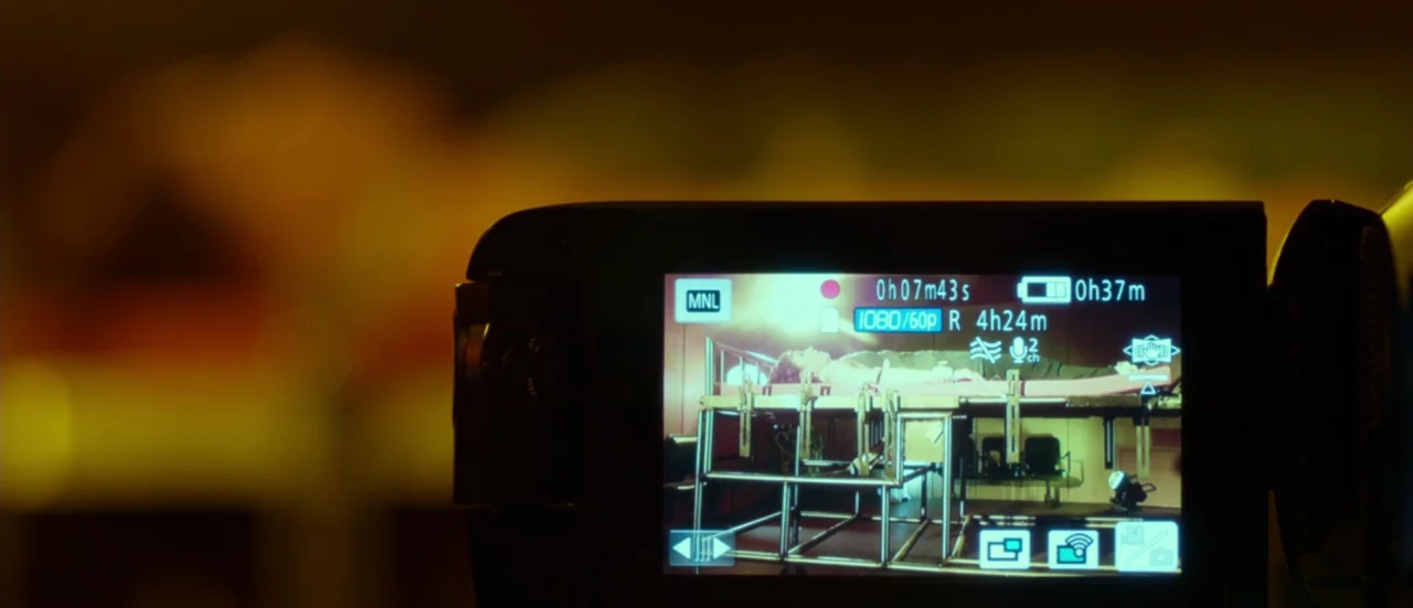 Rupture (2016) movie still: closeup — A digital camcorder screen displays a person lying on a metal examination bed. The room contains industrial…