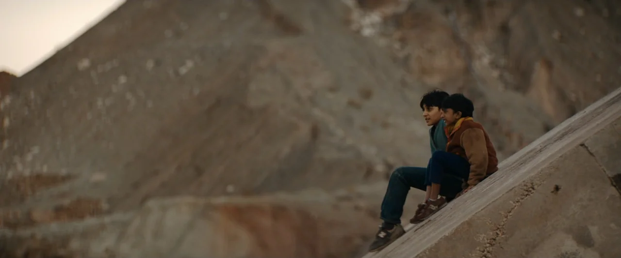Encounter (2018) movie still: wide — Two children sit on a concrete slope set against a large, barren rock formation. The boys are positioned…