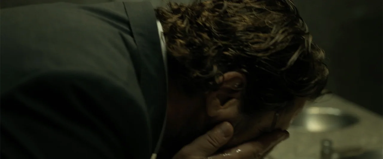 Den of Thieves 2: Pantera (2025) movie still: closeup — A dimly lit room with stone textures and a metallic sink basin. A man in a dark blazer leans…