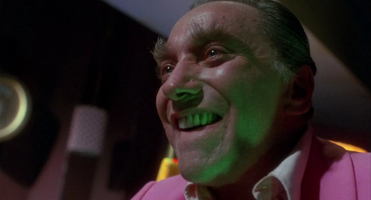 Vamp (1986) movie still: closeup — A dimly lit indoor space with vibrant colored lighting reflected on surfaces. An older man in a pink jacket…
