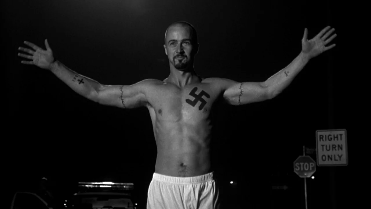 American History X movie still: fullbody — A night scene at a road junction featuring a single illuminated male figure. The man stands shirtless…
