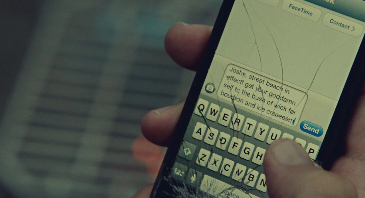 While We’re Young (2014) movie still: closeup — A close-up view shows a handheld smartphone with a shattered glass screen. A text message is being…