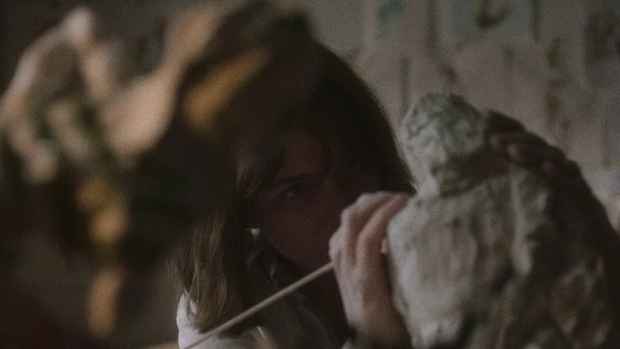 Showing Up (2022) movie still: medium — A dimly lit art studio space features a clay bust in the foreground. A woman focuses intently while using a…