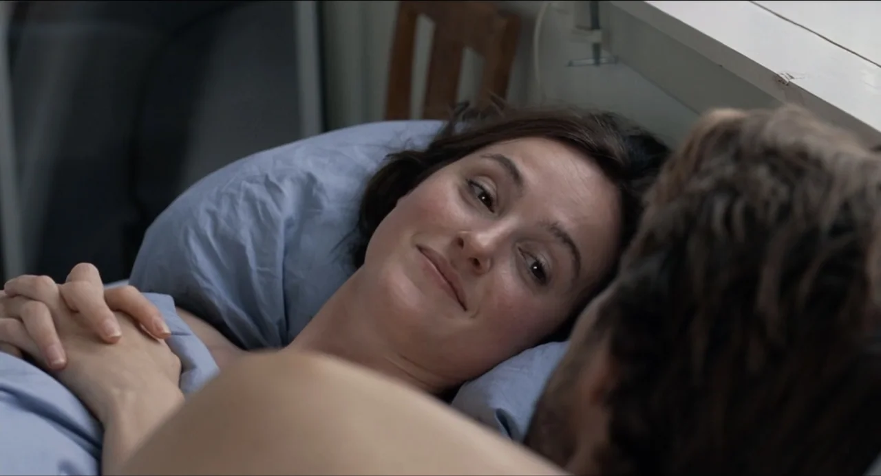 Sentimental Value (2025) movie still: closeup — A light-filled bedroom with a window visible in the background and blue bedding. A woman rests her…