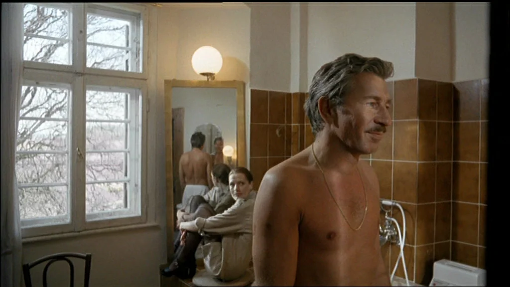 Chinese Roulette (1976) movie still: medium — A tiled room features a large mirror reflecting a woman sitting near a window. A shirtless man in the…