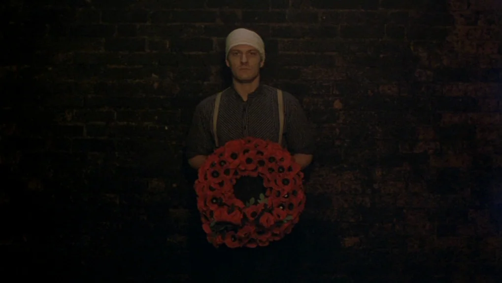 War Requiem (1989) movie still: medium — A man stands centered against a dark, textured brick wall in a dimly lit room. He holds a large circular…