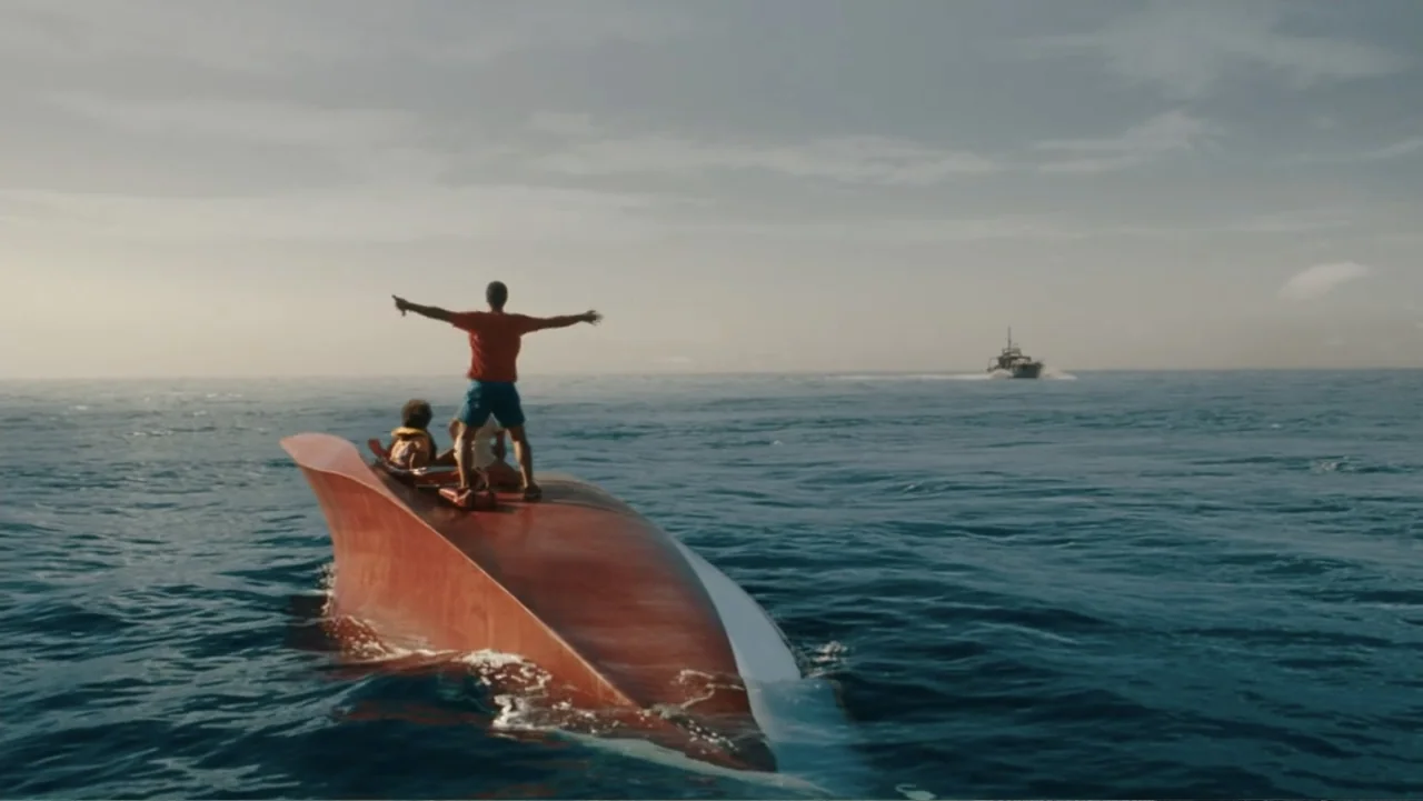 Jurassic World Rebirth (2025) movie still: wide — An overturned boat hull floats on calm open ocean water under an overcast sky. A man stands on top…