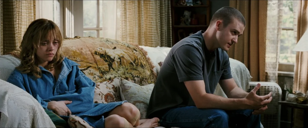 Black Snake Moan (2006) movie still: medium — A living room features a patterned sofa with a throw blanket and nearby window curtains. A woman in a…