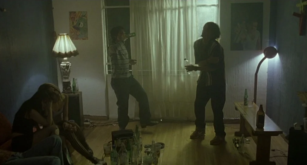 Y Tu Mamá También (2001) movie still: wide — An apartment interior is dimly lit by a lamp and outside light filtered through curtains. Three young…