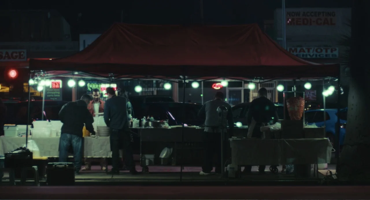 Good Fortune (2025) movie still: wide — A food stand with a red canopy occupies a parking lot at night. Several men prepare and serve food to…