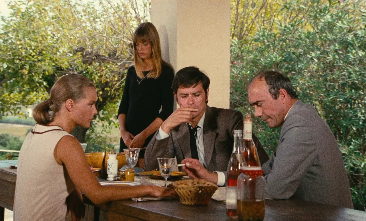 The Swimming Pool (1969) movie still: medium — Four people sit at a long wooden table on a bright, foliage-filled outdoor terrace. They are gathered…