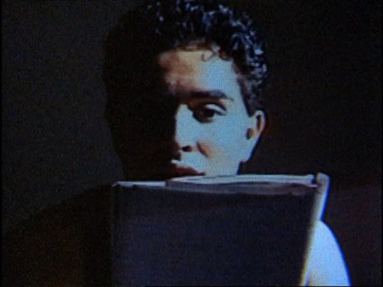 Totally F***ed Up (1993) movie still: closeup — A man stands in a dimly lit, dark space with a rough, low-resolution texture. He holds a rectangular…