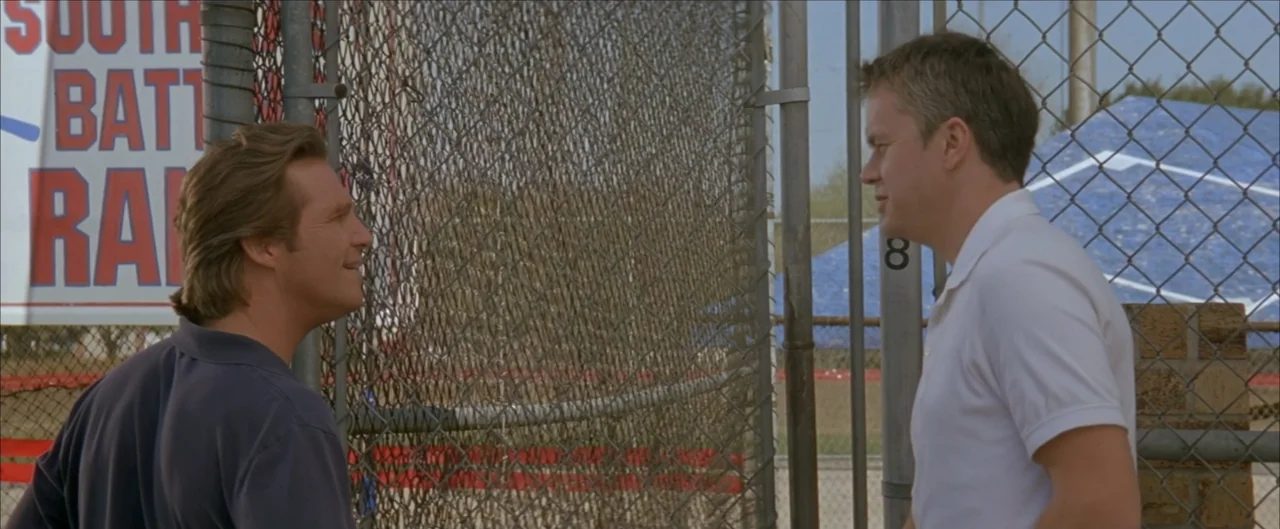Arlington Road (1999) movie still: medium — A baseball dugout exterior features a chain-link fence separating the two men. Two men in polo shirts…