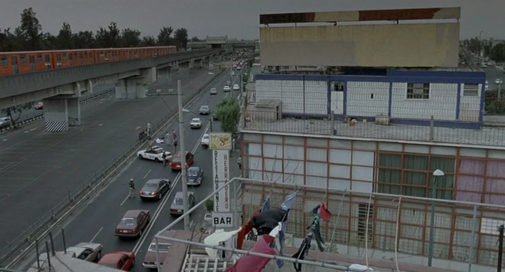 Y Tu Mamá También (2001) movie still: establishing — A desolate urban landscape features a multi-lane highway running parallel to an elevated train…