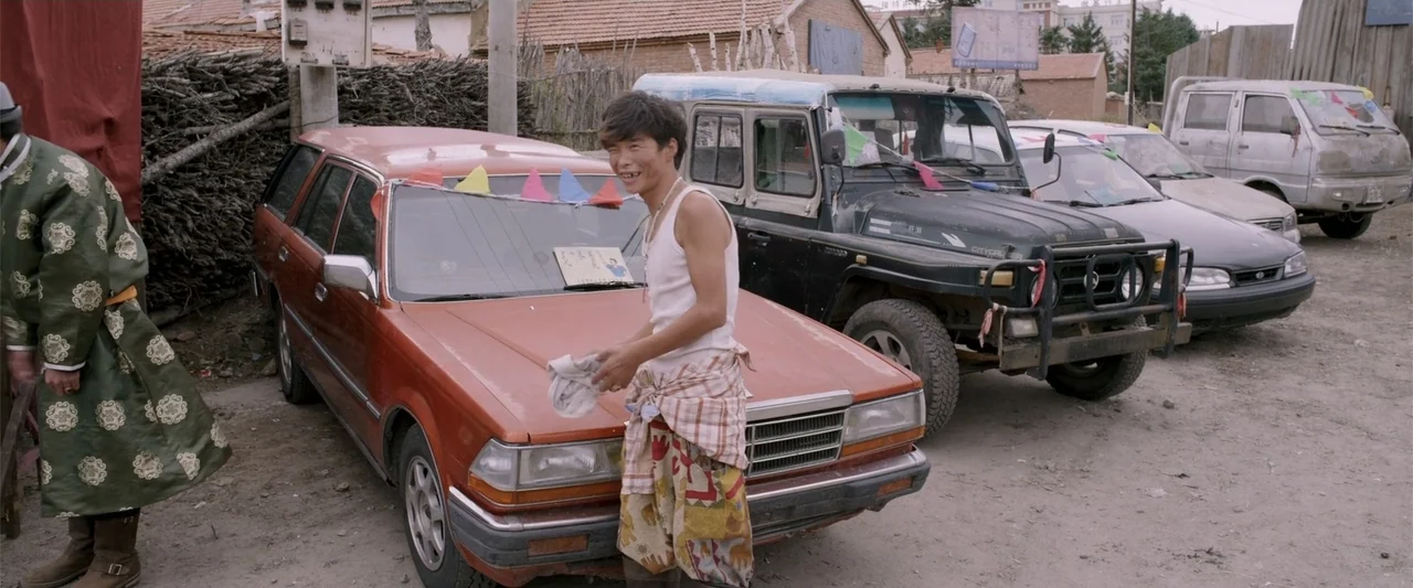 Skiptrace (2016) movie still: fullbody — An outdoor lot contains several parked vehicles including a red station wagon. A young man stands by the…