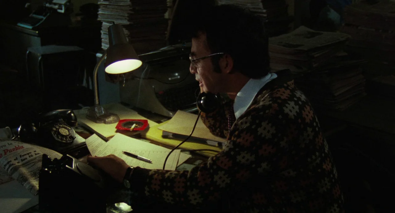 Canoa: A Shameful Memory (1976) movie still: medium — A dark, cluttered room filled with stacks of paper and vintage equipment. A man sits at a desk…