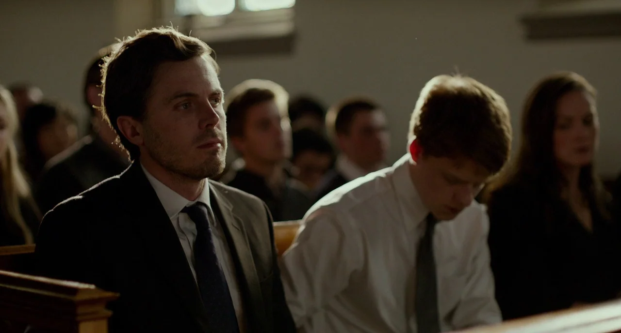 Manchester By The Sea (2016) movie still: medium — A group of people sit in rows of wooden pews inside a building. Two men in formal attire occupy…