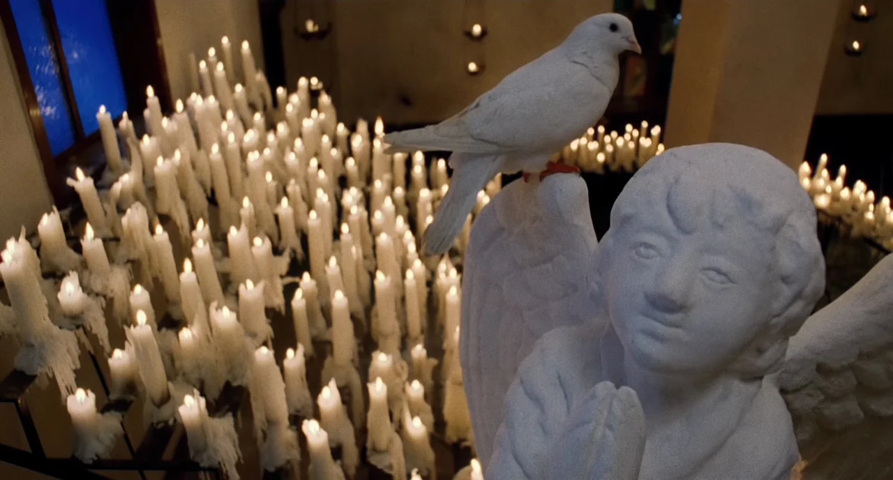 The Killer (1989) movie still: medium — A stone angel statue with hands in prayer sits surrounded by hundreds of candles. A white dove rests upon…