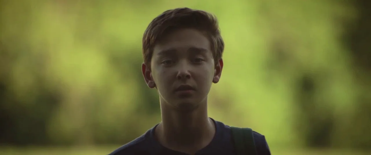 The Strange Ones (2017) movie still: closeup — A teenage boy stands in a blurred outdoor area with green trees. He looks toward the camera with a…