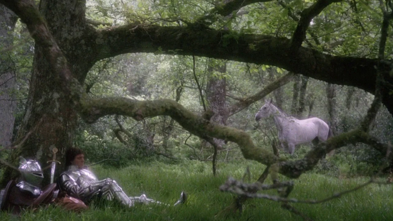 Excalibur (1981) movie still: wide — A green woodland with large trees and dense low-lying foliage. A knight wearing shiny plate armor leans against…
