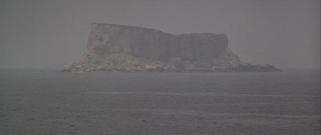 Popeye (1980) movie still: establishing — A flat-topped rocky island rises out of a calm, grey sea under an overcast sky. The distant landform is…