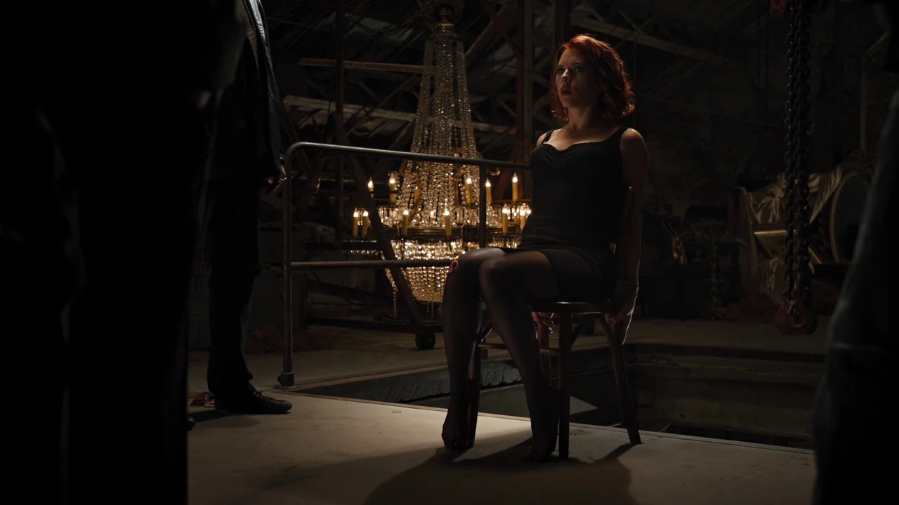 The Avengers (2012) movie still: medium — A dark warehouse interior featuring a large lit crystal chandelier in the background. A woman in a black…
