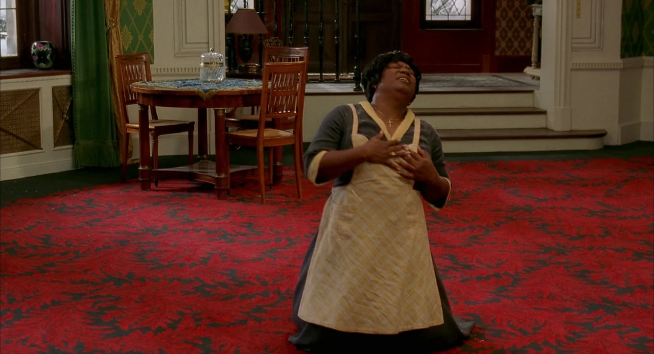 8 Women (2002) movie still: fullbody — A spacious room features a vibrant red patterned carpet and a formal table set. An middle-aged woman in a…