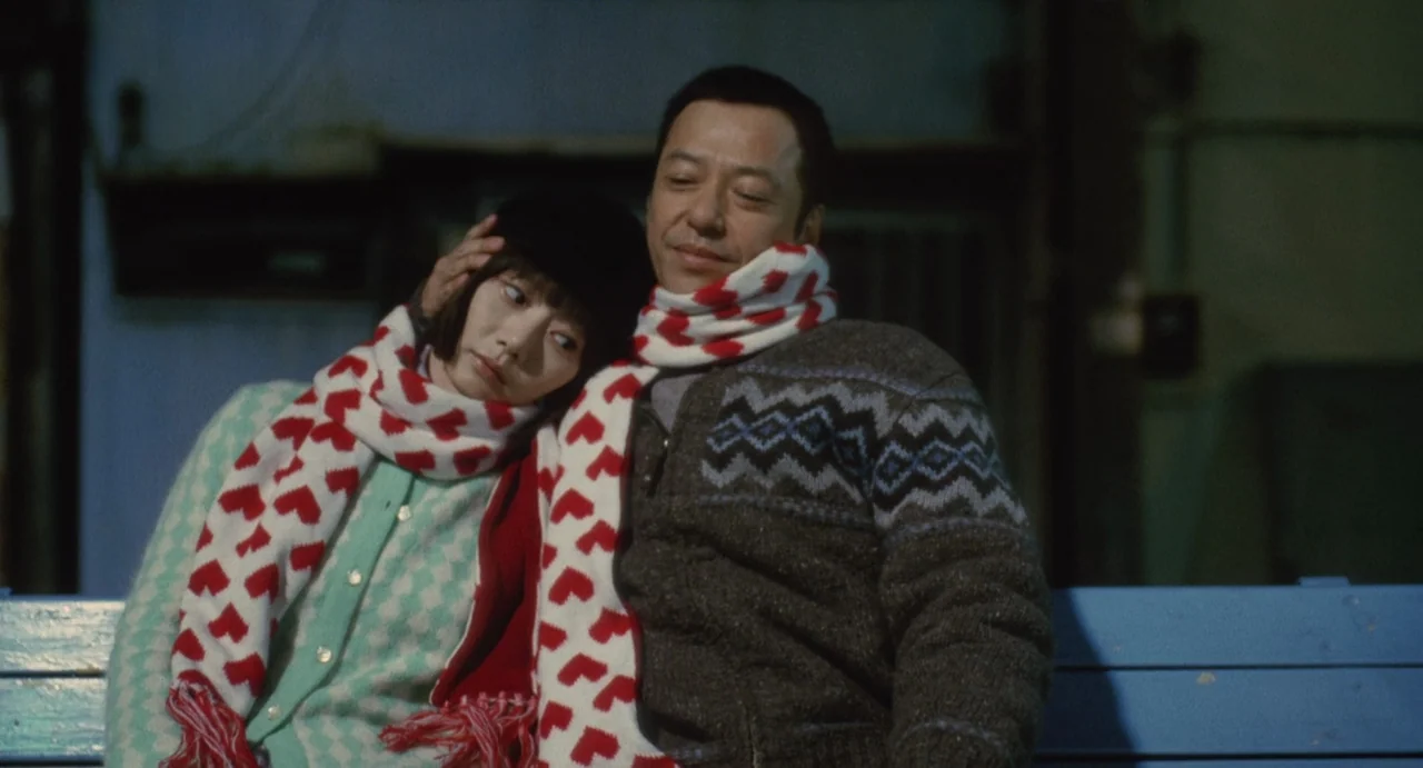 Air Doll (2009) movie still: medium — A man and woman sit together on a blue metal bench outdoors at night. The man rests his hand on the woman's…