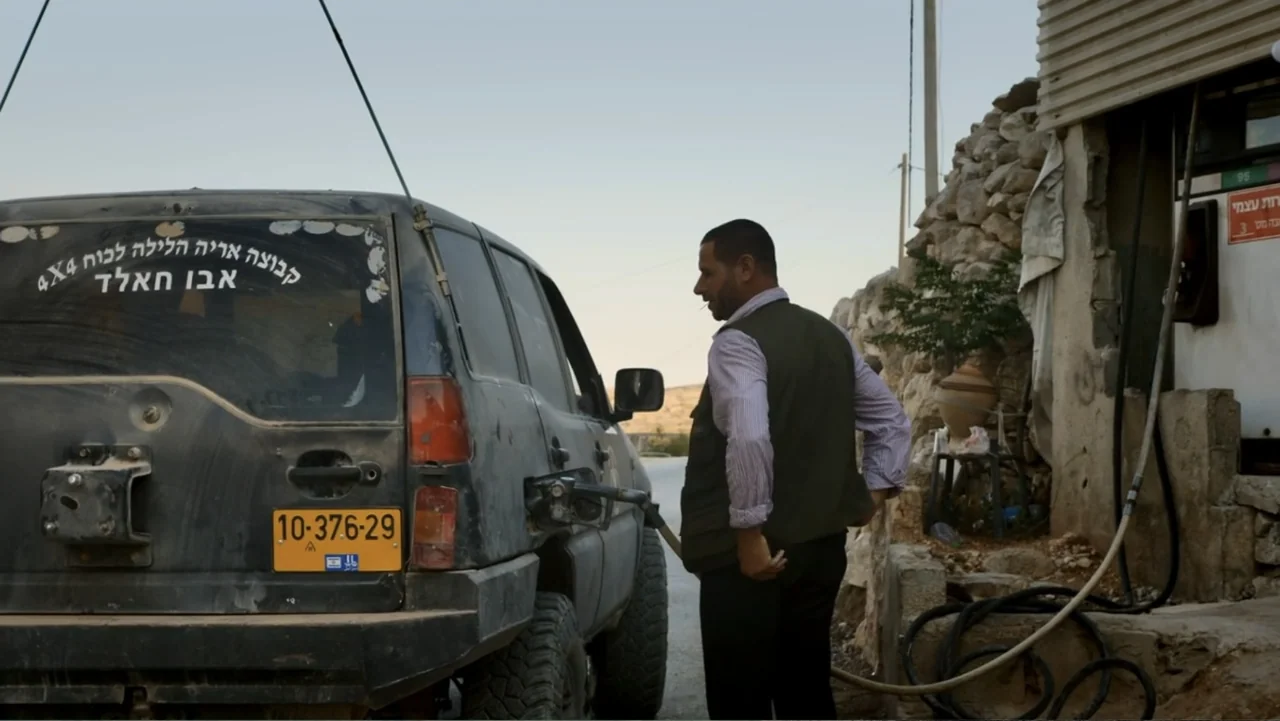 No Other Land (2024) movie still: medium — A rural gas station sits against a backdrop of rough stone masonry and dry hills. A man in an olive vest…
