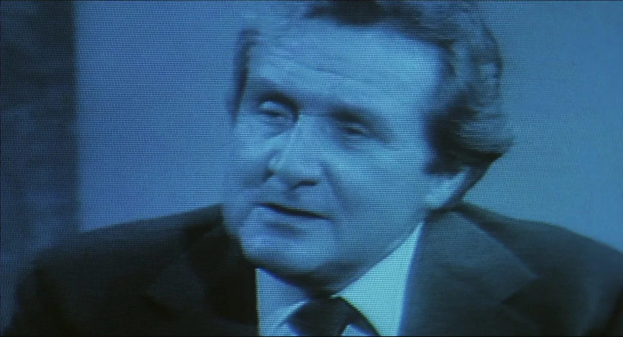 The Howling (1981) movie still: closeup — A man appears in a low-resolution broadcast transmission against a solid blue backdrop. He speaks while…