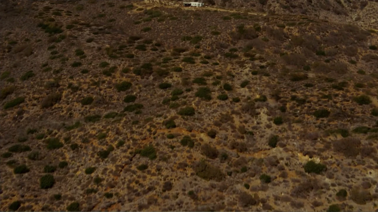 Hancock (2008) movie still: establishing — An arid hillside covered in sparse dry brush and dirt under bright sunlight. A white recreational vehicle…